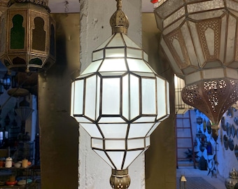 May include: A hanging, geometric-shaped lantern with a white glass body and brass-colored metal trim. The lantern is suspended from a chain and has a decorative finial at the top and bottom. Other ornate lanterns are visible in the background.
