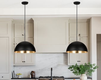 Può includere: Due lampade a sospensione nere con interno dorato sono appese al soffitto di una cucina. Le lampade hanno una forma a cupola e sono sospese da un cavo nero. La cucina presenta mobili bianchi e una combinazione di colori neutri.