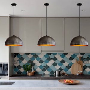 May include: Three black pendant lights with hammered metal shades hang over a kitchen counter with a blue and green tiled backsplash. The lights are turned on and emit a warm glow.