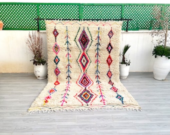 May include: A white Moroccan rug with a colorful diamond pattern. The rug has a fringe border and is made of wool.