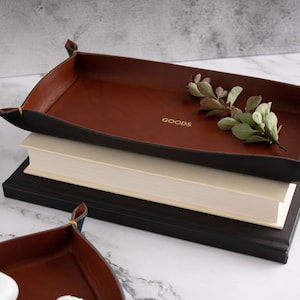 May include: A brown leather tray with black edges and gold stitching. The tray is sitting on top of two books. The tray has the word "GOODS" embossed in gold on the bottom.