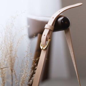 May include: A light beige leather strap with a gold metal buckle. The strap is attached to a wooden chair.