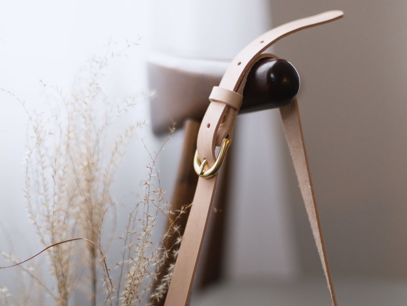 May include: A light beige leather strap with a gold metal buckle. The strap is attached to a wooden chair.