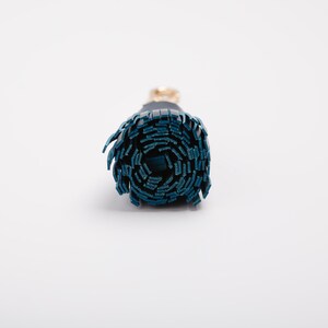 May include: A rolled-up, cylindrical object with a dark teal exterior and a gold-colored accent. The object appears to be made of a series of layered, rectangular pieces. The background is plain white.