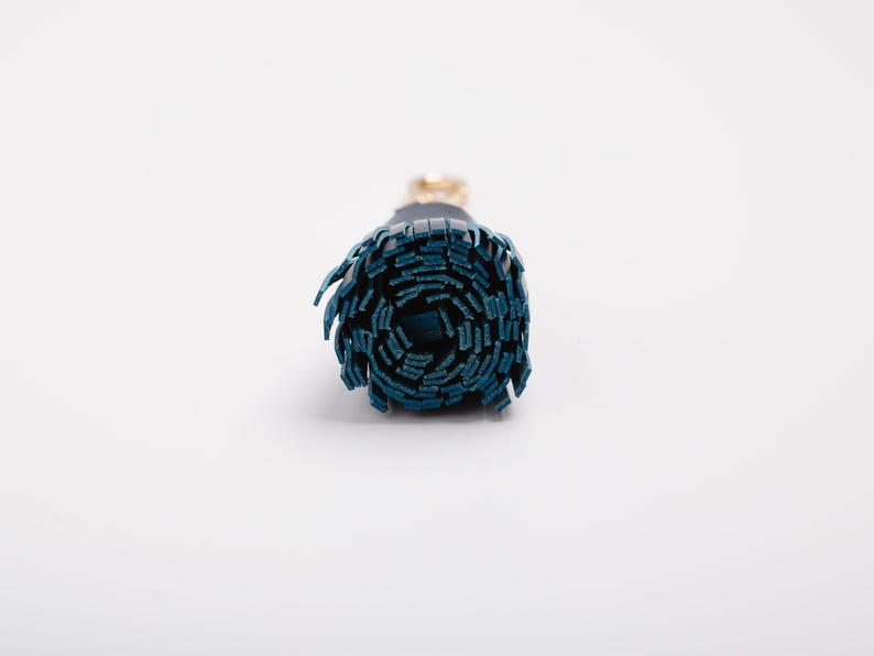 May include: A rolled-up, cylindrical object with a dark teal exterior and a gold-colored accent. The object appears to be made of a series of layered, rectangular pieces. The background is plain white.