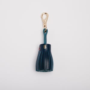May include: A navy blue leather tassel keychain with a gold-colored clasp and teal loop. The tassel has a gold letter "B" and red stitching. The keychain is on a white background.