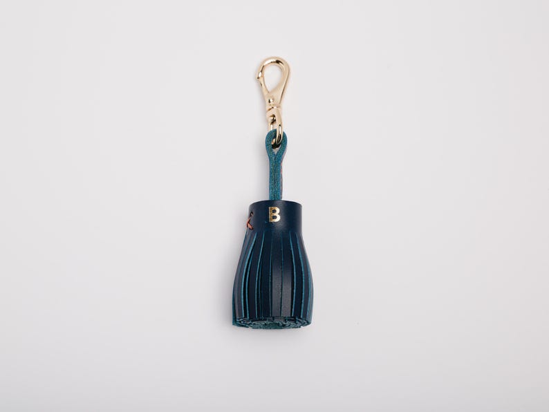 May include: A navy blue leather tassel keychain with a gold-colored clasp and teal loop. The tassel has a gold letter "B" and red stitching. The keychain is on a white background.