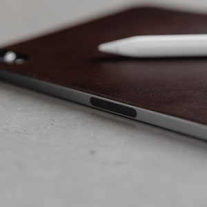 May include: A brown leather case for a tablet with a silver edge and a black power button. A white stylus is lying on top of the case.