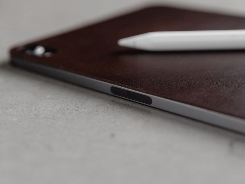 May include: A brown leather case for a tablet with a silver edge and a black power button. A white stylus is lying on top of the case.