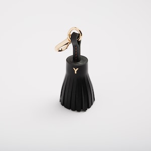 May include: Black leather tassel keychain with gold-tone hardware. The keychain features a gold-colored clasp and a black leather tassel with the letter "Y" in gold. The tassel is made of multiple strips of leather.