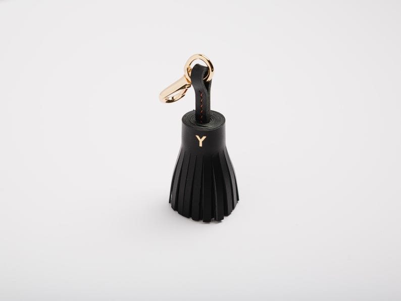 May include: Black leather tassel keychain with gold-tone hardware. The keychain features a gold-colored clasp and a black leather tassel with the letter "Y" in gold. The tassel is made of multiple strips of leather.
