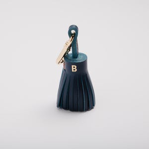 May include: A dark teal leather tassel keychain with a gold-colored clasp. The tassel has a gold-colored letter "B" on the front. The keychain is on a white background.