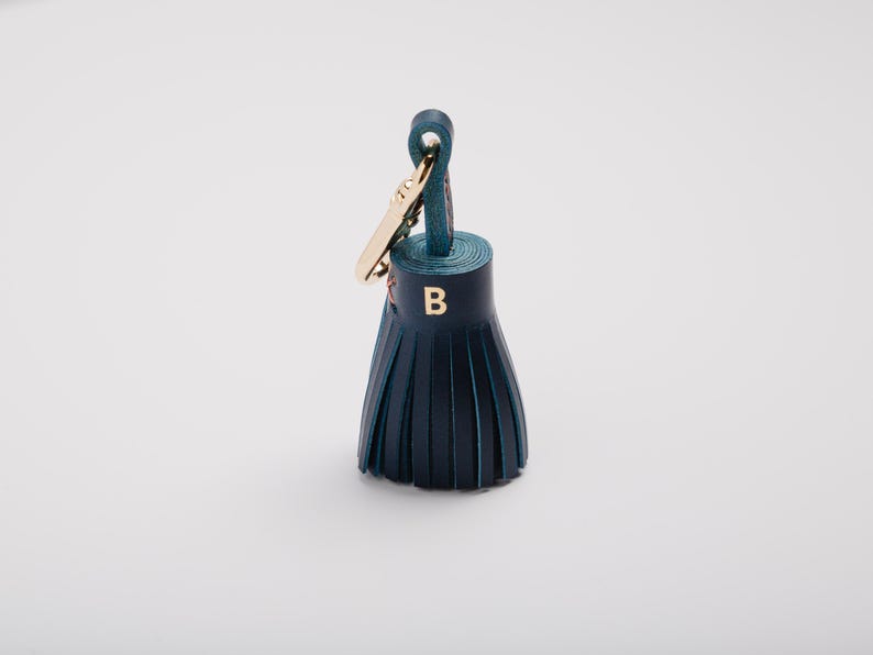 May include: A dark teal leather tassel keychain with a gold-colored clasp. The tassel has a gold-colored letter "B" on the front. The keychain is on a white background.
