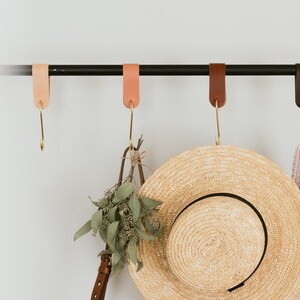 Puede incluir: Una barra de metal negro con cuatro correas de cuero y ganchos dorados que cuelgan de ella. Un sombrero de paja, una correa de cuero marrón con un montón de hojas verdes y un suéter rosa cuelgan de los ganchos.
