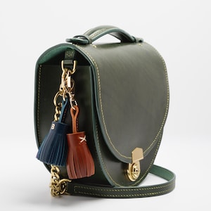 May include: A green leather handbag with a top handle and a gold chain strap. The bag features a gold clasp and decorative tassels in navy blue and brown. The bag is approximately 9 inches wide.