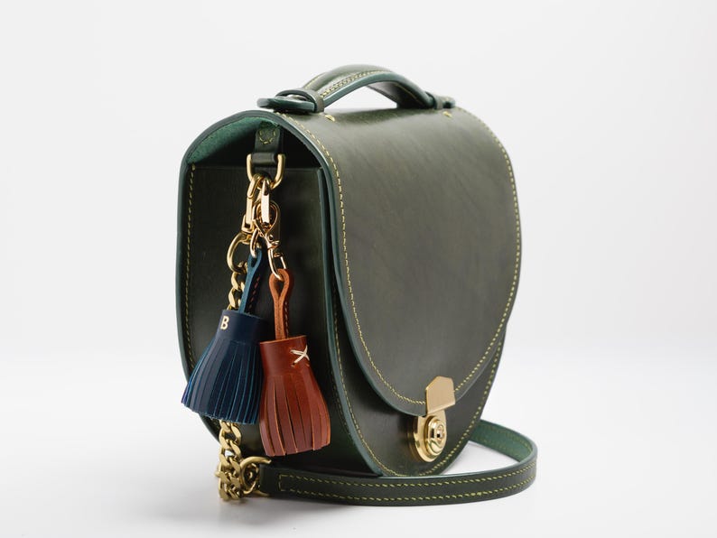 May include: A green leather handbag with a top handle and a gold chain strap. The bag features a gold clasp and decorative tassels in navy blue and brown. The bag is approximately 9 inches wide.