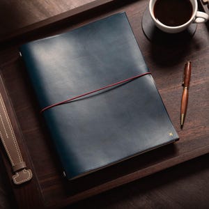 May include: A dark blue leather journal with a red elastic closure, resting on a wooden tray. A cup of coffee and a wooden pen are also on the tray. The journal has a small gold initial on the cover.