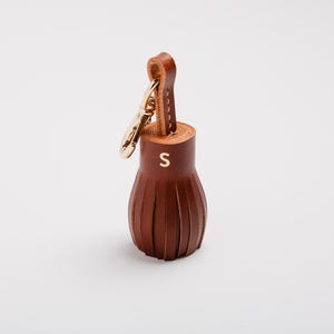 May include: A brown leather tassel keychain with a gold-colored clasp and loop. The tassel has a cylindrical top with the letter "S" in gold and a fringed bottom. The keychain is on a white background.