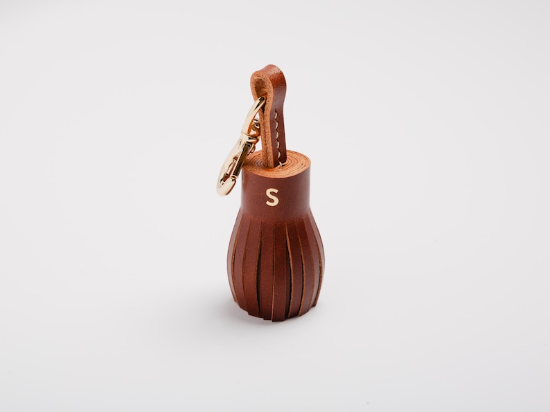 May include: A brown leather tassel keychain with a gold-colored clasp and loop. The tassel has a cylindrical top with the letter "S" in gold and a fringed bottom. The keychain is on a white background.