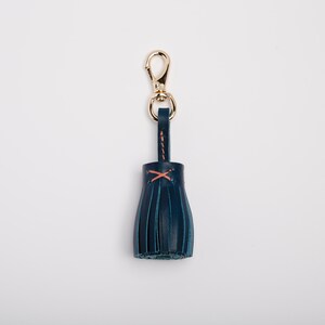 May include: A navy blue leather tassel keychain with a gold-colored clasp. The tassel is stitched with orange thread. The keychain is a stylish accessory.