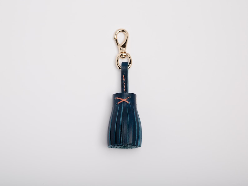 May include: A navy blue leather tassel keychain with a gold-colored clasp. The tassel is stitched with orange thread. The keychain is a stylish accessory.
