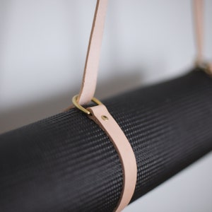May include: A black yoga mat with a light brown leather strap and brass hardware. The strap is attached to the mat with two brass rings.