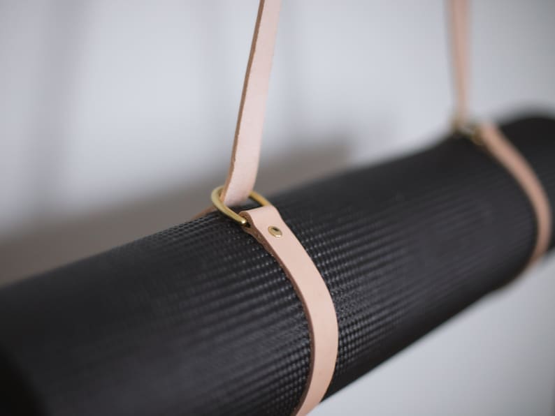 May include: A black yoga mat with a light brown leather strap and brass hardware. The strap is attached to the mat with two brass rings.