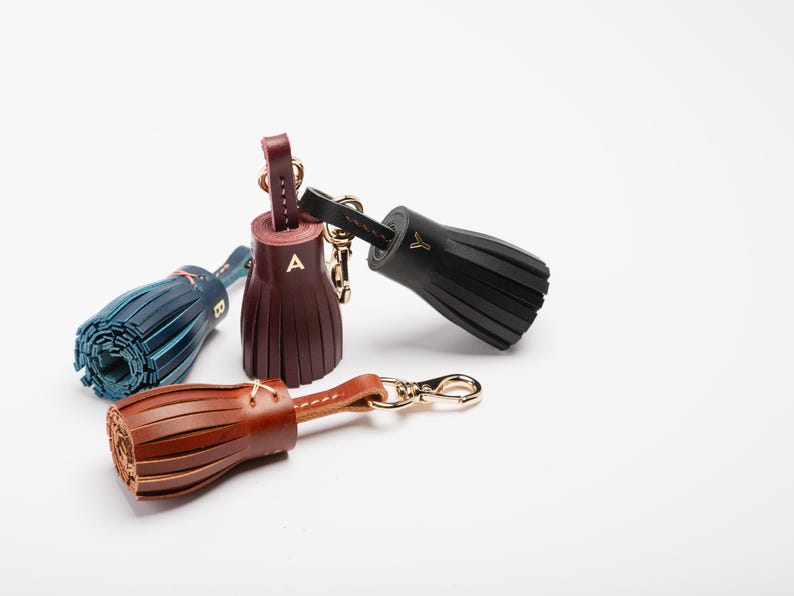 May include: Leather tassel keychains in brown, black, burgundy, and teal blue. Each keychain features a gold-colored clasp and a leather loop. The burgundy tassel has the letter "A" and the teal blue tassel has the letter "B".