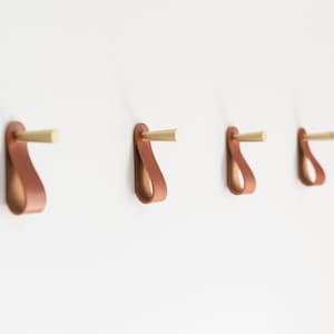 May include: Four brown leather hooks with brass accents mounted on a white wall.