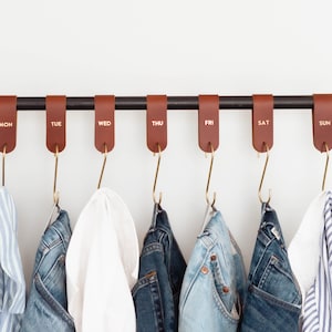 May include: A brown leather hanging organiser with gold hooks labelled with the days of the week. The organiser is hanging on a black rod and has various clothing items hanging from the hooks.