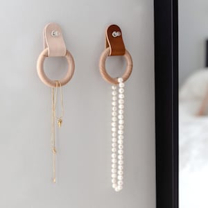 Puede incluir: Dos anillos de madera con correas de cuero colgando de una pared blanca. Un collar de oro con un colgante de corazón cuelga de un anillo, y un collar de perlas blancas cuelga del otro.