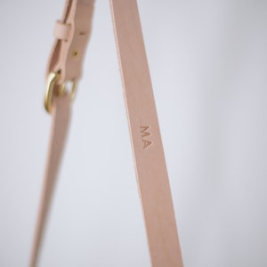 May include: Close-up of a light brown leather strap with a brass buckle and the initials "MA" embossed on the strap.
