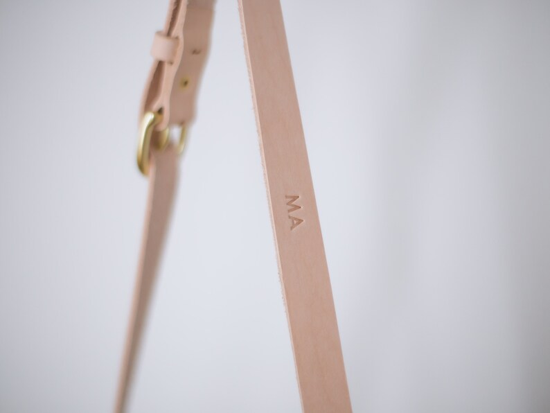 May include: Close-up of a light brown leather strap with a brass buckle and the initials "MA" embossed on the strap.