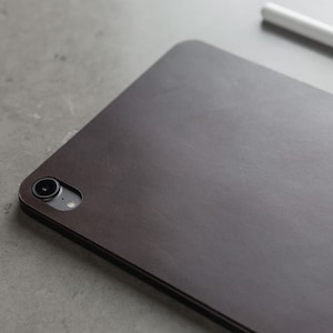 May include: A dark brown leather case for a tablet or e-reader. The case has a camera lens cutout and a subtle textured surface.