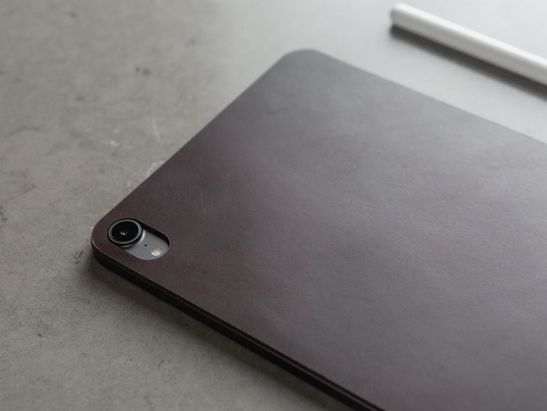 May include: A dark brown leather case for a tablet or e-reader. The case has a camera lens cutout and a subtle textured surface.