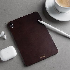 May include: A dark brown leather case for a tablet with the initials "JDM" embossed in gold on the bottom right corner. A white stylus and a white case for wireless earbuds are also in the image.
