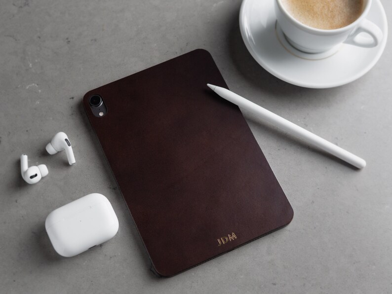 May include: A dark brown leather case for a tablet with the initials "JDM" embossed in gold on the bottom right corner. A white stylus and a white case for wireless earbuds are also in the image.