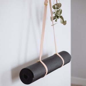 May include: A rolled-up black yoga mat hanging from a light brown leather strap with gold hardware. The strap is attached to a white wall.