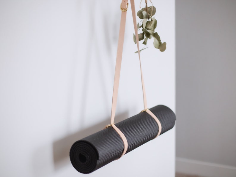 May include: A rolled-up black yoga mat hanging from a light brown leather strap with gold hardware. The strap is attached to a white wall.