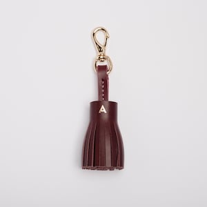 May include: A burgundy leather tassel keychain with a gold-colored clasp and a small gold letter "A" on the tassel. The tassel is made of vertical strips of leather, and the keychain is against a white background.