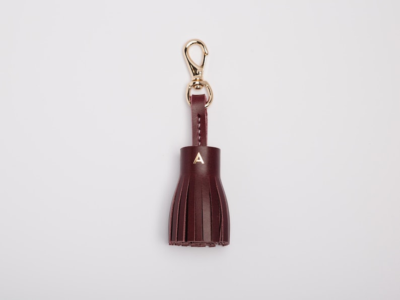 May include: A burgundy leather tassel keychain with a gold-colored clasp and a small gold letter "A" on the tassel. The tassel is made of vertical strips of leather, and the keychain is against a white background.