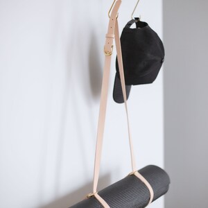 May include: A black yoga mat hanging from a metal rack with a light brown leather strap. A black baseball cap hangs from a hook on the rack.