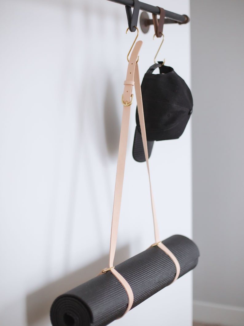 May include: A black yoga mat hanging from a metal rack with a light brown leather strap. A black baseball cap hangs from a hook on the rack.