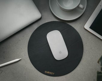 Op de afbeelding: Een ronde, zwarte leren muismat met het woord "CHRISSY" in gouden letters. Een witte draadloze muis ligt op de mat.