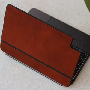 Puede incluir: Una funda de cuero marrón para portátil con un portátil negro en su interior. La funda tiene una solapa que se pliega sobre el portátil y se cierra con un cierre magnético.