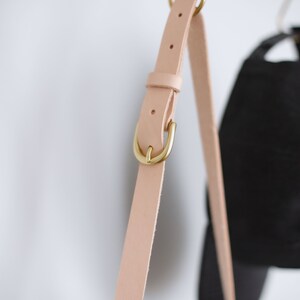 May include: A close-up of a light pink leather strap with a gold metal buckle. The strap is hanging from a hook.