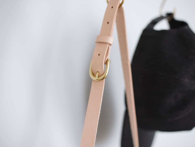 May include: A close-up of a light pink leather strap with a gold metal buckle. The strap is hanging from a hook.