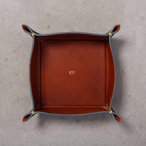 May include: A brown leather tray with blue stitching and yellow accents. The tray has a square shape and is monogrammed with the initials "KF".