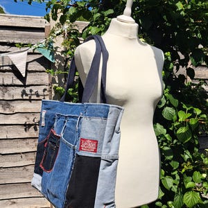 May include: A patchwork denim tote bag constructed from recycled jeans, showcasing different denim washes. The bag has navy blue straps and a red Superdry label. The bag is displayed on a mannequin outdoors, surrounded by greenery.