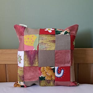 May include: A square patchwork cushion with a variety of fabric squares in shades of red, pink, brown, and yellow. Some squares feature floral and geometric patterns. The cushion is on a white surface.